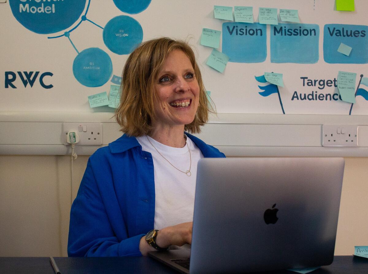 A coach smiling at her laptop in front of a wall of Vision to Value strategy notes